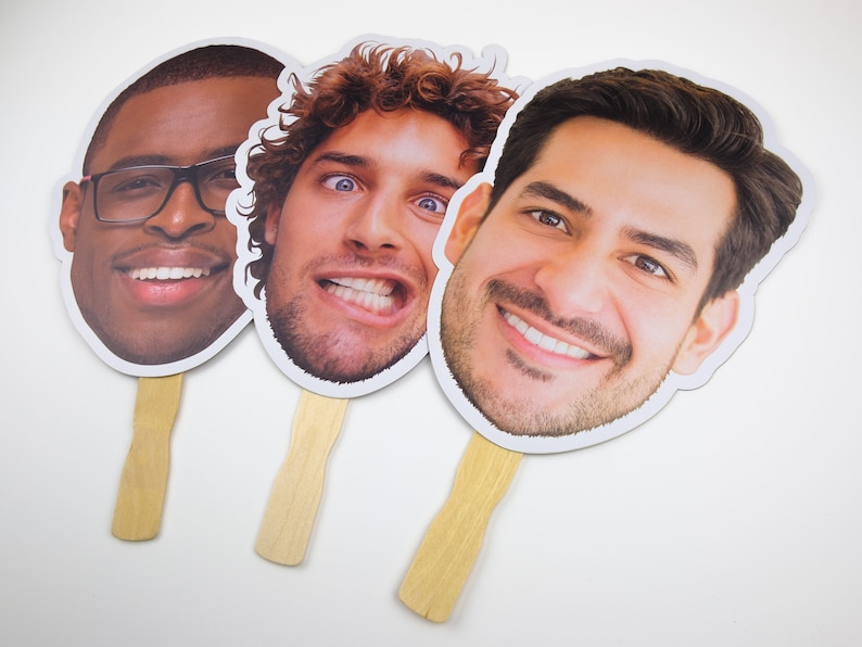 May include: Three cardboard face cutouts on sticks. The faces are of different people with different expressions. The cutouts are white with a white border.