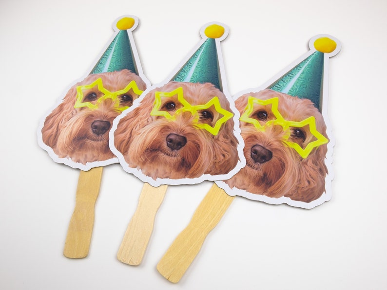 May include: Three party favor fans featuring a dog wearing a green party hat and yellow star-shaped glasses. The fans have wooden sticks.