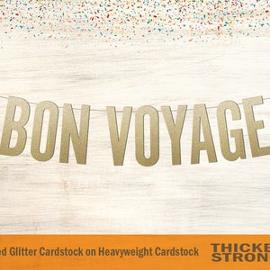 Peut inclure: Banni&egrave;re en carton paillet&eacute; dor&eacute; avec l'inscription "BON VOYAGE" suspendue &agrave; une ficelle sur un fond de bois blanc. La banni&egrave;re est en carton &eacute;pais et ne perd pas ses paillettes.