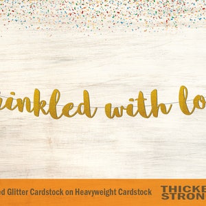 Peut inclure: Banni&egrave;re dor&eacute;e &agrave; paillettes avec le texte "sprinkled with love" sur un fond en bois blanc. La banni&egrave;re est faite de papier cartonn&eacute; paillet&eacute; non-effiloch&eacute; sur papier cartonn&eacute; &eacute;pais.