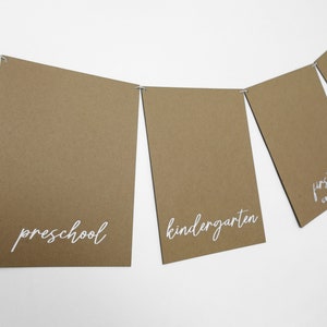 May include: A banner of brown rectangular cards strung together with wire. Each card has white cursive text indicating school grades: preschool, kindergarten, and first grade. The background is plain white.