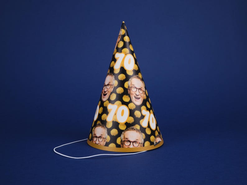 Peut inclure: Un chapeau de f&ecirc;te noir et or avec une ficelle blanche. Le chapeau est d&eacute;cor&eacute; d'un motif de points jaunes et du nombre "70" r&eacute;p&eacute;t&eacute; plusieurs fois. Il y a aussi des images de visage de personne sur le chapeau.