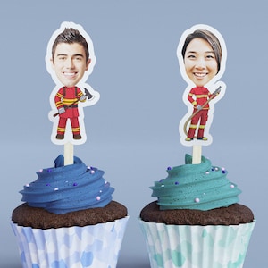 May include: Two cupcakes with blue and green frosting and sprinkles. Each cupcake has a topper with a cartoon image of a person dressed as a firefighter.