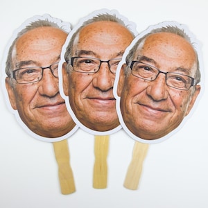 May include: Three white cardboard face masks with a photo of an older man wearing glasses. The masks are attached to wooden sticks.