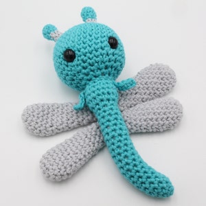 May include: A crocheted dragonfly toy with a teal body and gray wings. The dragonfly has black eyes and a white antenna.