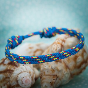 Op de afbeelding: Een gevlochten armband van blauw, geel en rood touw met een knoop aan het ene uiteinde. De armband ligt op drie schelpen.