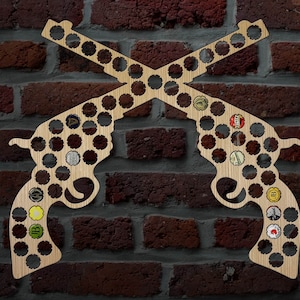 May include: Wooden wall art shaped like crossed revolvers, designed to hold bottle caps. The art piece is made of light-colored wood and is mounted on a brick wall. The bottle caps are visible within the gun silhouettes, adding color and detail.