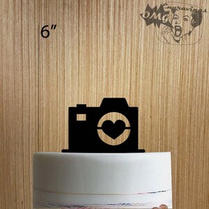 May include: Black camera-shaped cake topper with a heart-shaped lens, measuring 6 inches. The topper is placed on a white cake. The background is a light brown wood grain. The image also includes the text "OMG Engravings".