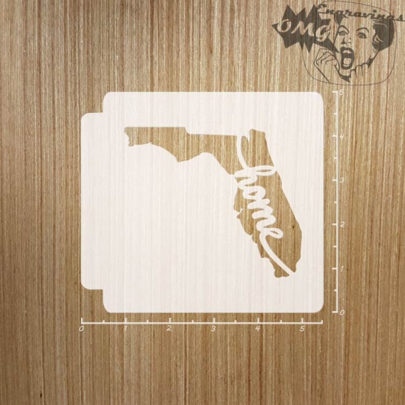 May include: A white stencil featuring the state of Florida with the word "home" in a cursive font. The stencil is on a wooden surface. A logo is in the upper right corner. The stencil measures approximately 5 inches.