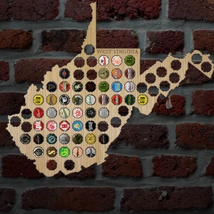 May include: Wooden West Virginia state-shaped wall art with holes to hold beer bottle caps. The sign has the words "WEST VIRGINIA" at the top. The background is a brick wall.