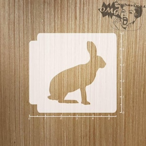 May include: A white stencil of a rabbit silhouette, measuring 5 inches wide and 4 inches tall. The stencil is on a wooden surface.
