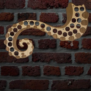 May include: A wooden cat-shaped wall decoration with a swirling tail design. The cat is made of light brown wood with circular cutouts and a decorative pattern. The background is a brick wall.