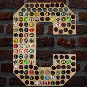 May include: A wooden letter C with a brown wood grain finish. The letter is decorated with bottle caps of various colors and designs. The letter is mounted on a brick wall.