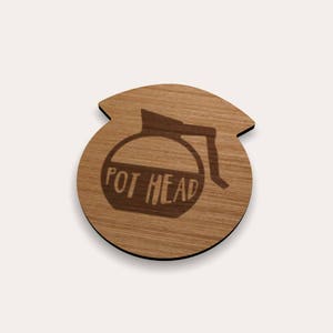 Op de afbeelding: Houten onderzetter in de vorm van een koffiepot met de woorden "POT HEAD" erin gegraveerd. De onderzetter is gemaakt van lichtbruin hout en heeft een gladde afwerking. Het ontwerp is een humoristische kijk op een koffiepot.
