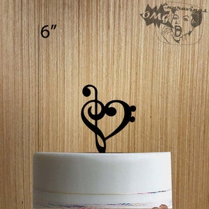 Puede incluir: Un adorno para tarta negro en forma de corazón con una clave de sol y una clave de fa, de 15 cm. El adorno está colocado encima de una tarta blanca. El fondo es una superficie de madera.