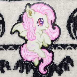 May include: A sticker featuring a cartoon pony with pink hair, wings, and a tail. The pony has a light green body and a red and purple eye. The sticker is set against a white and black Halloween-themed background.