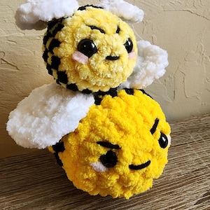 May include: A handmade plush bee toy with yellow and black stripes, white wings, and a smiling face. The bee has large black eyes and pink cheeks. The toy is made of soft, textured yarn and is designed to look like a cartoon bee.
