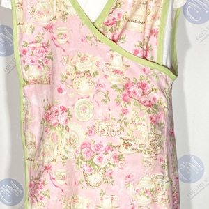 May include: Pink apron with a floral and teacup pattern, trimmed with green binding. The apron features a wrap-around design and is made of a soft fabric. The pattern includes roses, teacups, and teapots.