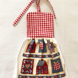 May include: A kitchen towel with a red and white gingham top and a hanging loop. The bottom has a patchwork design with houses, hearts, and apples in red, blue, and beige. The towel is made of fabric.