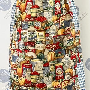 May include: Apron with a colorful print featuring apples, pumpkins, and various canned goods. The apron has a blue and white gingham pattern on the sides and top, with a blue border at the bottom. The design evokes a rustic, country theme.