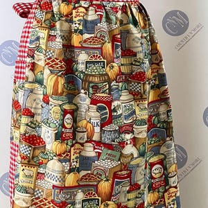 May include: A colorful apron with a red and white checkered waistband and ties. The apron's main fabric features a detailed print of various food items, including pumpkins, pies, and canned goods, creating a charming, vintage aesthetic.