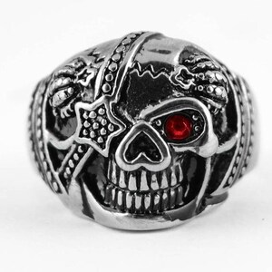 Könnte beinhalten: Ein silberner Totenkopfring mit einem roten Edelstein als Auge und einem sternförmigen Pflaster über dem anderen Auge. Der Totenkopf ist detailliert mit einer herzförmigen Nase und einem bandagierten Kopf.