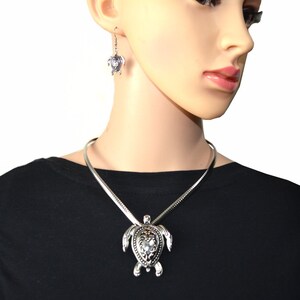 May include: Silver turtle necklace and earrings set. The necklace features a large turtle pendant with intricate details. The earrings are small and feature a turtle design.