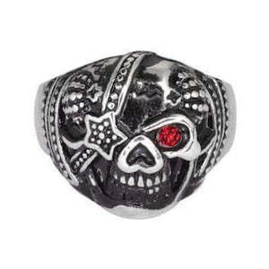 Peut inclure: Une bague en argent en forme de crâne avec un œil en pierre rouge et des détails complexes. Le crâne porte un bandana avec un motif d'étoile.
