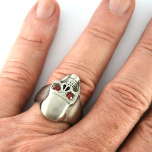 May include: A silver skull ring with red gemstone eyes. The ring is worn on a person's finger.