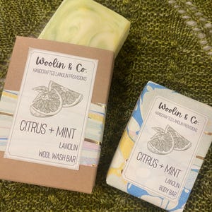 Puede incluir: Dos barras de jabón con aromas de cítricos y menta. El jabón de la izquierda es una barra de lavado de lana y el jabón de la derecha es una barra para el cuerpo. Ambos jabones tienen una etiqueta con una ilustración de cítricos y el texto "Woolin & Co. Handcrafted Lanolin Provisions Citrus + Mint Lanolin"