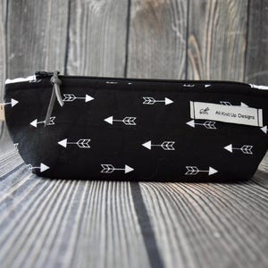 Puede incluir: Estuche negro con estampado de flechas blancas. El estuche rectangular tiene cierre de cremallera y una pequeña etiqueta con una escala de medición. El texto "All Knit Up Designs" está en una etiqueta blanca.