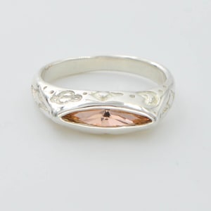 May include: A silver ring with an oval, peach-colored gemstone set in the center. The band features a decorative pattern. The ring is displayed on a white background.