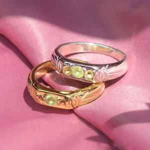 May include: Two rings with green gemstones and intricate floral designs. The top ring is silver and the bottom ring is gold.