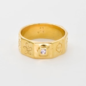 May include: A gold-colored ring with a square, light purple gemstone set in the center. The ring has a textured surface with etched floral and other decorative designs. The ring is on a white background.