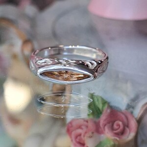 May include: A silver ring with an oval, light brown gemstone. The ring band has a textured design. The ring is displayed on a reflective surface with blurred pink and floral elements in the background.