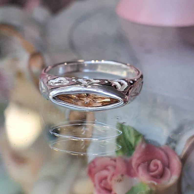 May include: A silver ring with an oval, light brown gemstone. The ring band has a textured design. The ring is displayed on a reflective surface with blurred pink and floral elements in the background.
