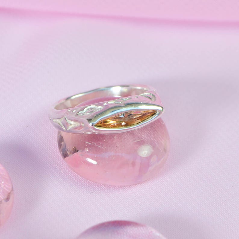 May include: A silver ring with an oval, amber-colored gemstone. The ring is set on a pink, rounded stone against a soft pink background. The ring's band features a decorative pattern.