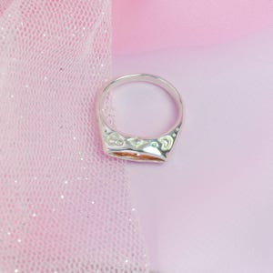 May include: A silver ring with a decorative design around the band. The ring is set against a pink background with a sheer, glittery fabric. The ring's design includes floral and geometric patterns.