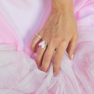 May include: Two silver rings are displayed on a hand against a pink background. One ring features a heart-shaped design with a pink and white pattern. The other ring has an orange gemstone. The background is a soft pink fabric.