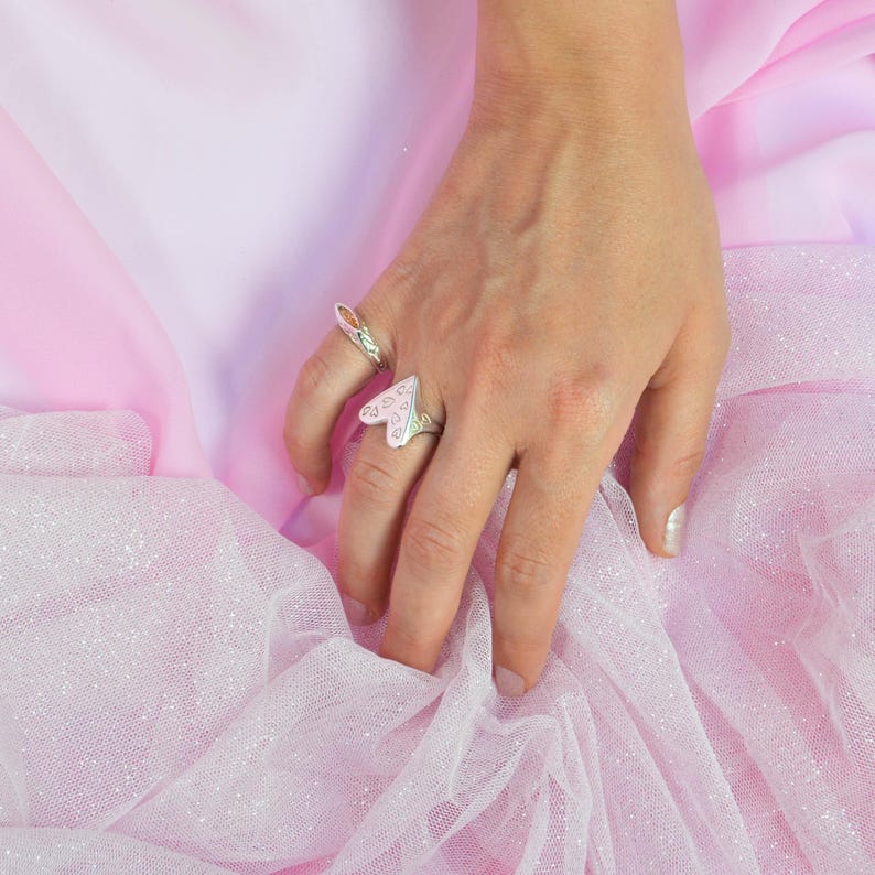 May include: Two silver rings are displayed on a hand against a pink background. One ring features a heart-shaped design with a pink and white pattern. The other ring has an orange gemstone. The background is a soft pink fabric.