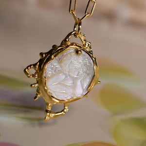 May include: A gold necklace with a white cameo pendant featuring a carved image of a woman's face. The pendant is surrounded by a gold frame with a textured design.