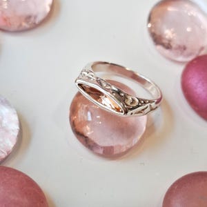 May include: A silver ring with an oval-shaped, light-orange gemstone. The ring has an ornate, engraved design along the band. The ring is resting on a pink, rounded stone, with other pink stones in the background.
