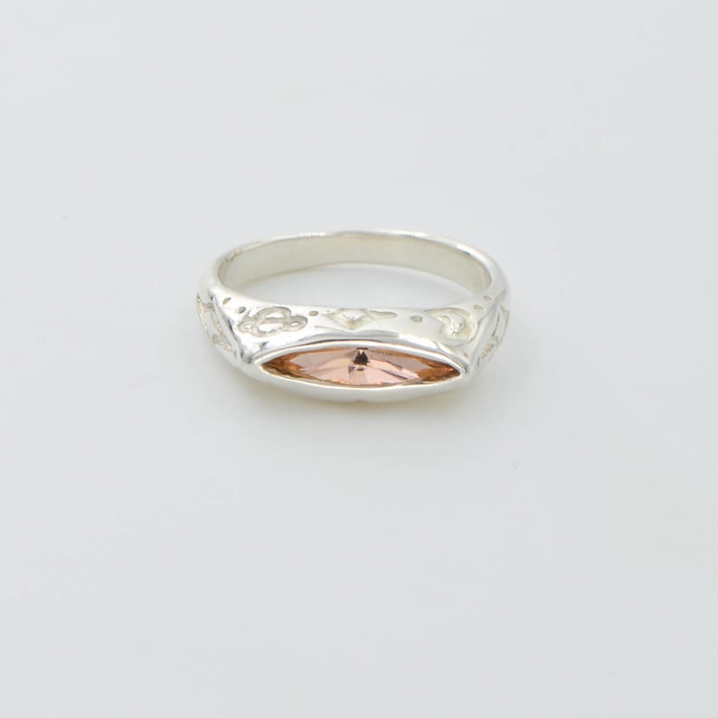 May include: A silver ring with an oval, peach-colored gemstone set in the center. The band features a decorative pattern. The ring is displayed on a white background.