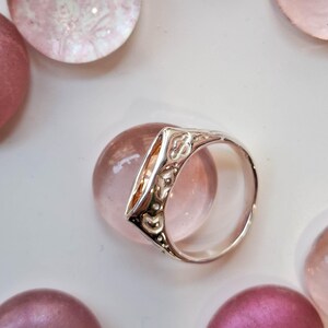 May include: A silver ring with an ornate design, featuring a triangular face with a clear, amber-colored stone. The ring is set against a backdrop of pink and purple glass spheres, creating a delicate and elegant aesthetic.