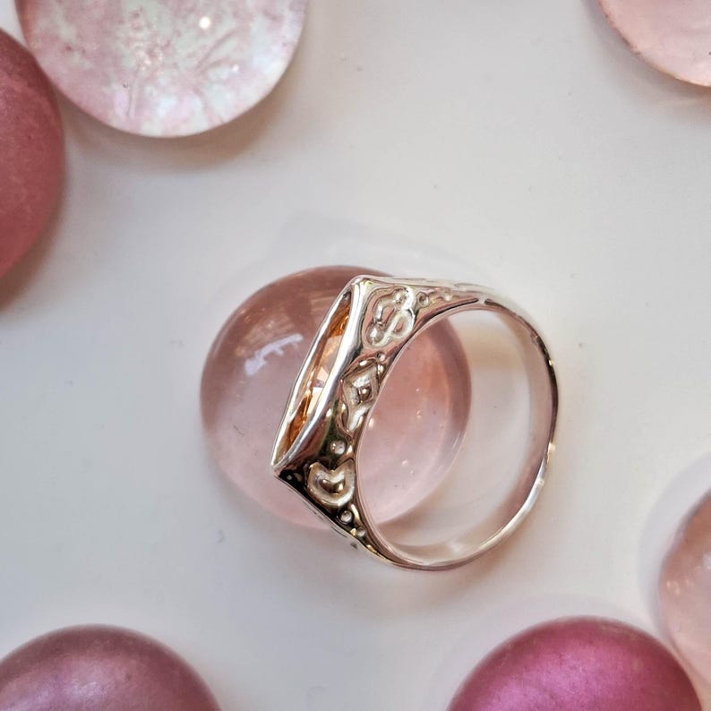 May include: A silver ring with an ornate design, featuring a triangular face with a clear, amber-colored stone. The ring is set against a backdrop of pink and purple glass spheres, creating a delicate and elegant aesthetic.