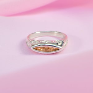 May include: A silver ring with an oval, amber-colored stone. The ring has a decorative design around the stone and on the band. The ring is set against a pink background.