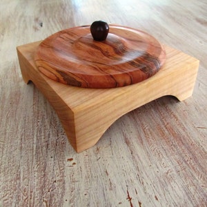 May include: A wooden box with a lid, the lid has a dark brown knob. The box is made of light wood and the lid is made of a darker wood with a grain pattern. The box is sitting on a light wood surface.