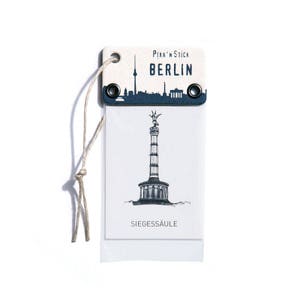 May include: A white tag with blue accents featuring a black and white illustration of the Siegessäule, a landmark in Berlin, Germany. The tag reads "Pikk'n'Stick Berlin" and "Siegessäule".
