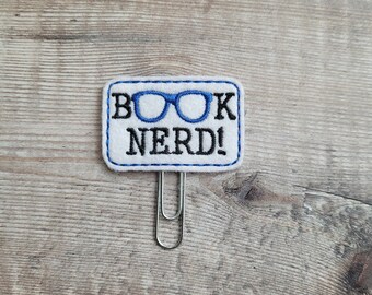 Op de afbeelding: Witte vilt boekenlegger met blauwe bril en de tekst "BOOK NERD!" in zwarte borduurwerk. De boekenlegger heeft een zilveren metalen clip.