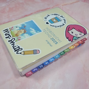 May include: A yellow and cream Hobonichi Techo 2025 planner with a clear plastic cover. The planner has several stickers on it, including a pencil, a coffee cup, a cartoon girl, and a sticker that says "Mrs. Smith". The planner has colorful tabs for each month of the year.